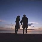Couple hold hands at sunset on a beach he is wearing shorts she is in a short dress. They are silhouetted against a blue sky.